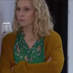 Kobieta z klanu obsada, w zielonej bluzce w kwiaty i musztardowym kardiganie, z rękami skrzyżowanymi na piersi.