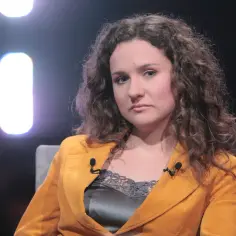 Kobieta z kręconymi włosami w musztardowej marynarce, być może kim jest żona Cejrowskiego, siedzi na kanapie.