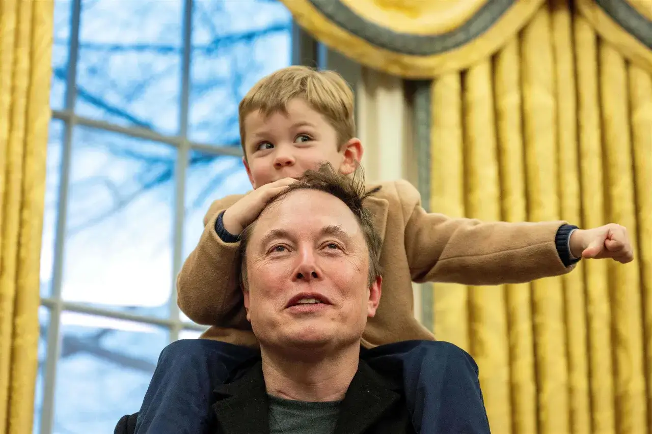 Uśmiechnięty Elon Musk z dzieckiem na ramionach. Chłopiec w beżowym płaszczyku wyciąga rękę, patrząc w bok.
