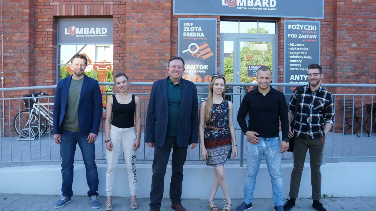 Lombard życie pod zastaw: obsada pozuje przed budynkiem. W tle szyldy z ofertą pożyczek i skupu złota.